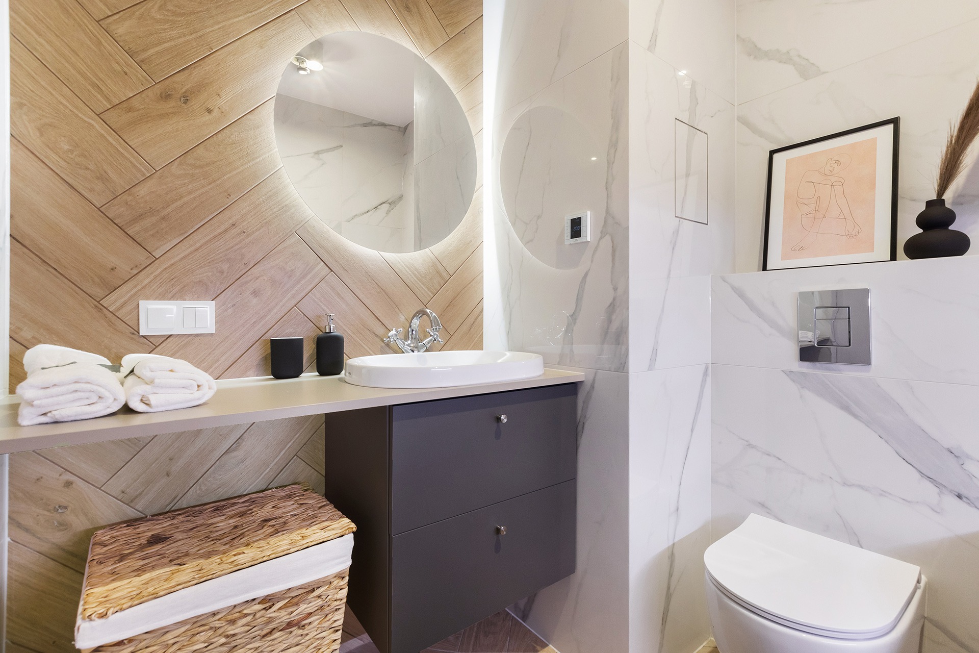 Nowoczesna łazienka z drewnianą ścianą ułożoną w jodełkę, okrągłym lustrem z podświetleniem LED, białą umywalką nablatową, szarą szafką pod umywalką i marmurowymi płytkami na ścianach