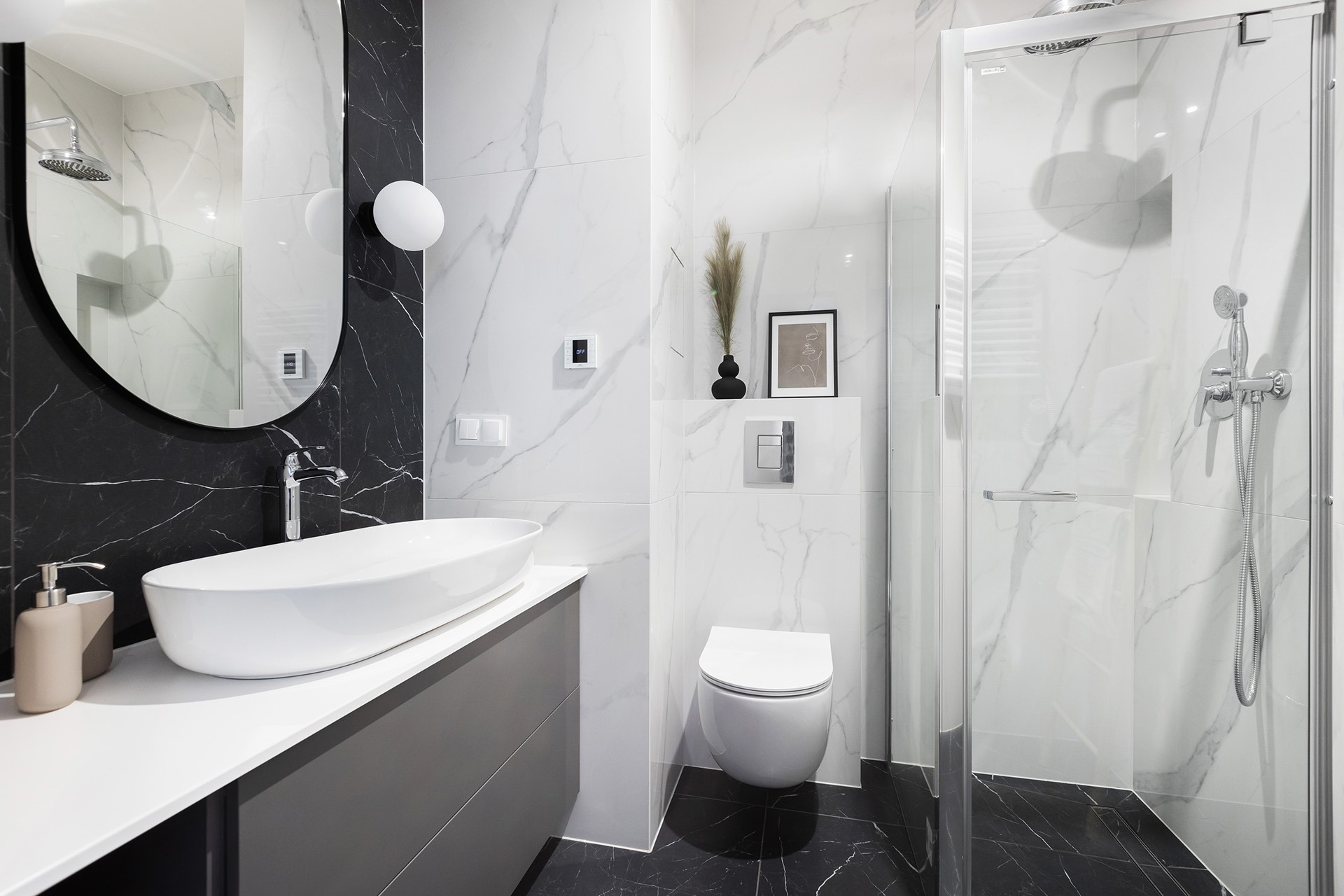 Nowoczesna łazienka z czarno-białymi marmurowymi płytkami, owalną umywalką nablatową, szarą szafką pod umywalką, kabiną prysznicową ze szklanymi drzwiami i wiszącą miską WC.