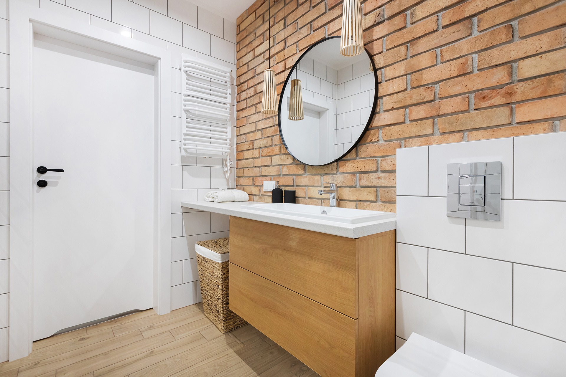 Nowoczesna łazienka z ceglaną ścianą, okrągłym lustrem, drewnianą szafką pod umywalką i białymi płytkami.