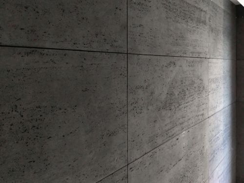 Ściana wykończona płytami imitującymi surowy beton, z widoczną teksturą i fugami, ułożonymi w regularny wzór.