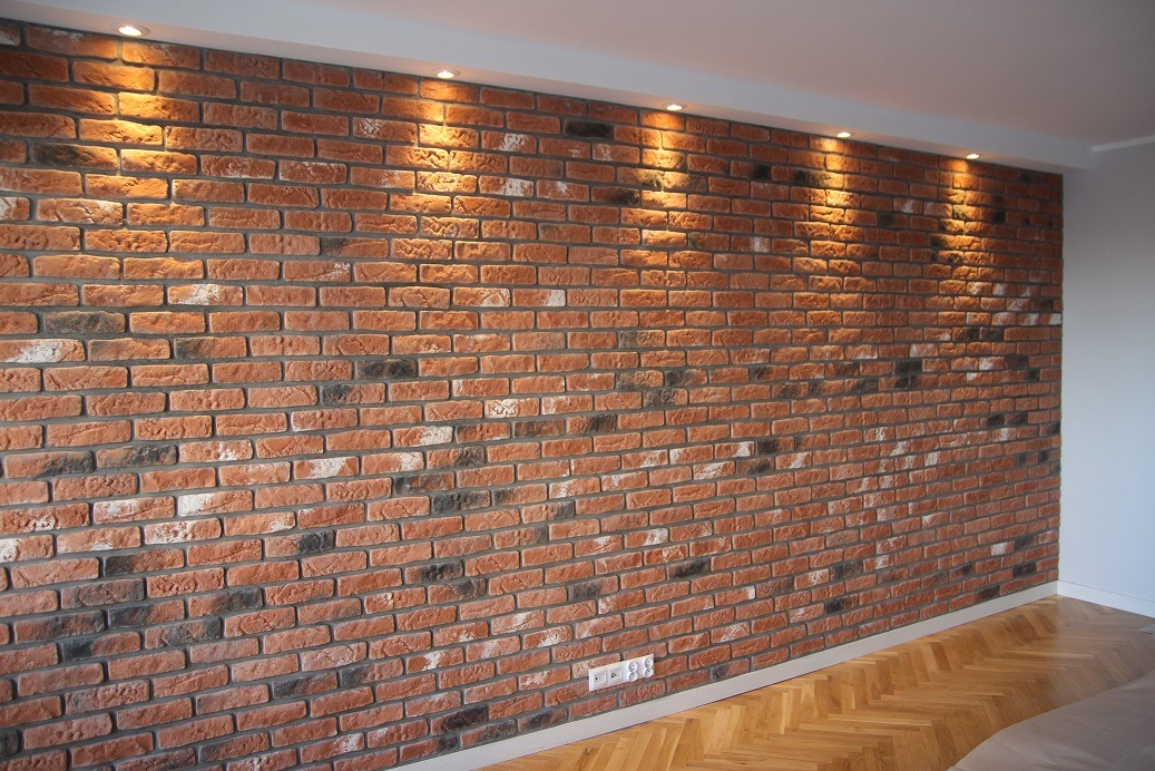 Ściana wykończona cegłą dekoracyjną w odcieniach czerwieni i szarości, oświetlona punktowymi lampami sufitowymi, z widocznym fragmentem parkietu u dołu i gniazdkiem elektrycznym przy listwie...