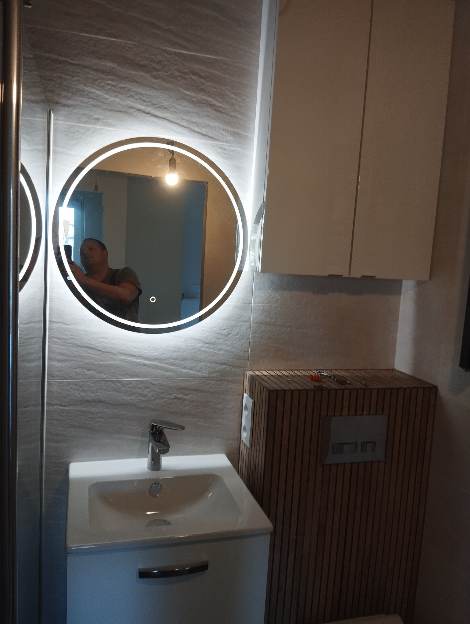 Nowoczesna łazienka w Pozowicach z okrągłym lustrem LED, białą szafką umywalkową i drewnianą obudową toalety. Minimalistyczny design i funkcjonalne rozwiązania.