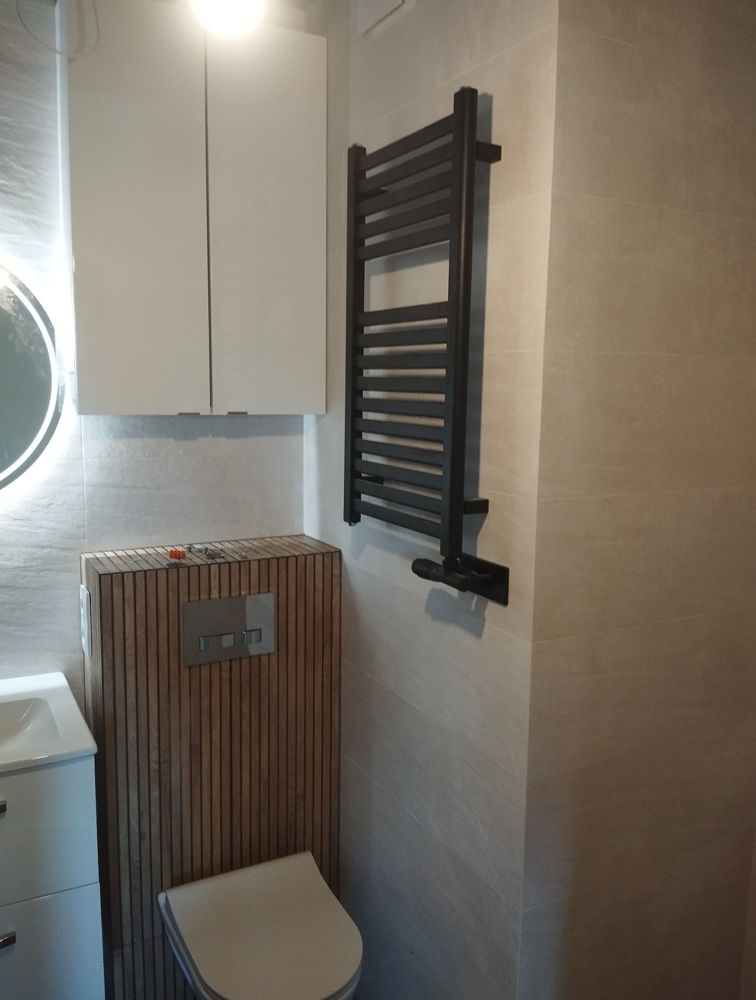 Nowoczesna łazienka z drewnianą obudową toalety, białymi szafkami i grzejnikiem drabinkowym. Jasne płytki na ścianach. Widoczne narzędzia na obudowie.