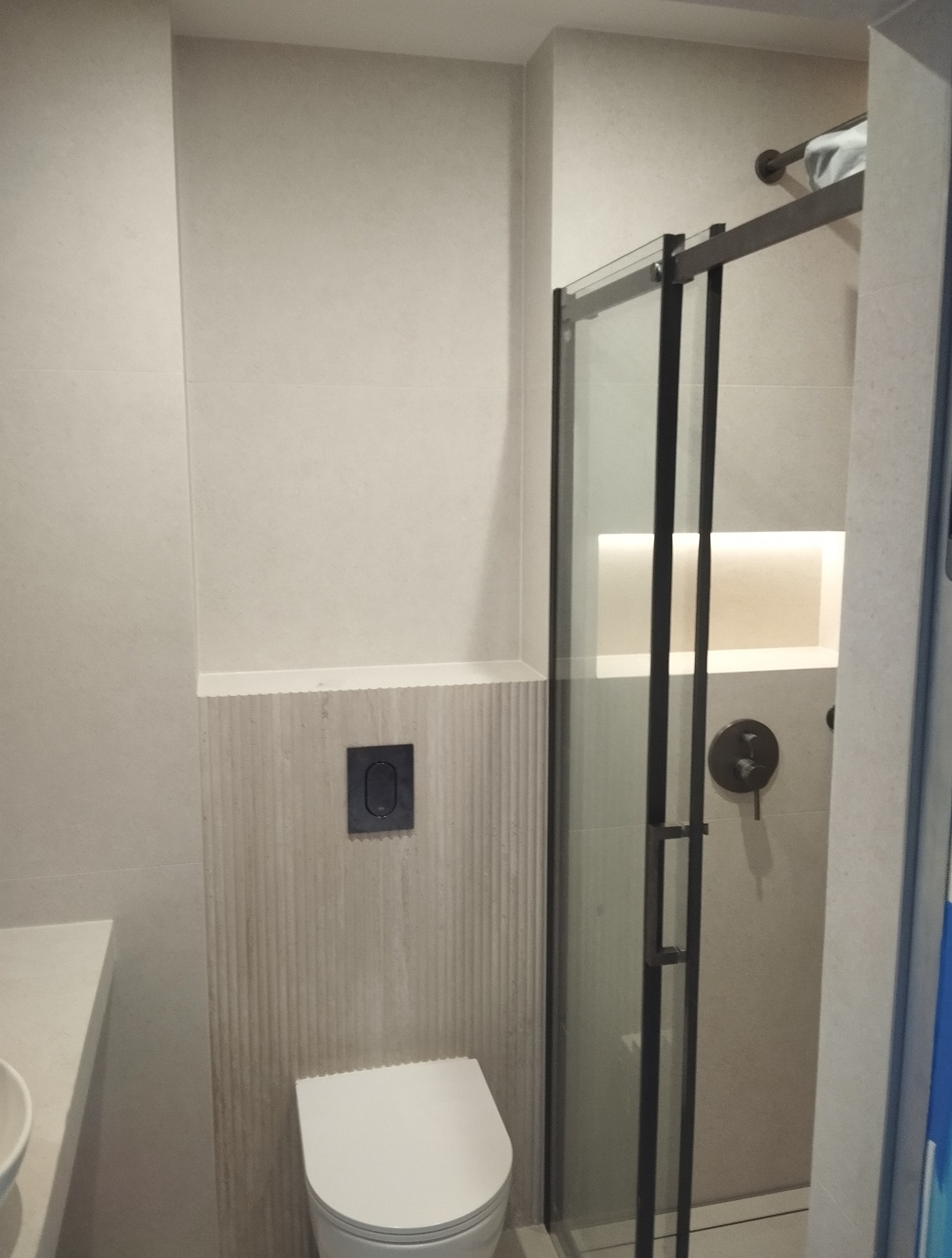 Nowoczesna łazienka z kabiną prysznicową typu walk-in, beżowe płytki z subtelną fakturą, podwieszana toaleta i minimalistyczny panel sterujący.