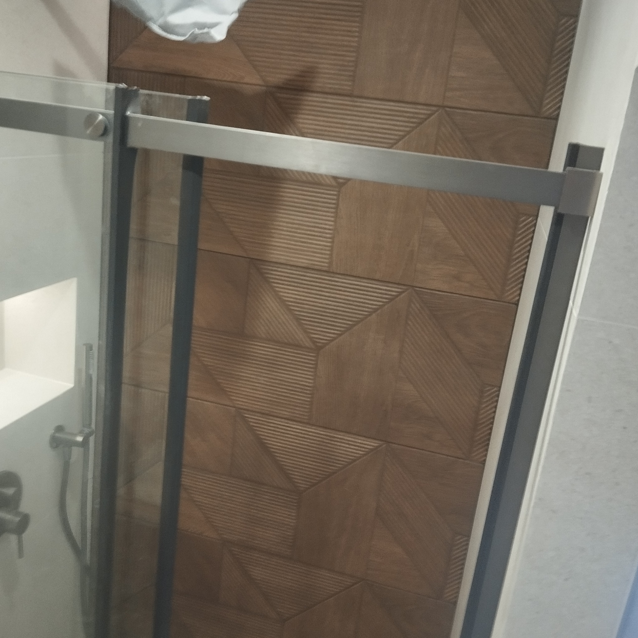 Nowoczesna kabina prysznicowa z geometrycznymi płytkami imitującymi drewno. Szklane drzwi z metalową ramą. Widoczna bateria i fragment półki wewnątrz kabiny.