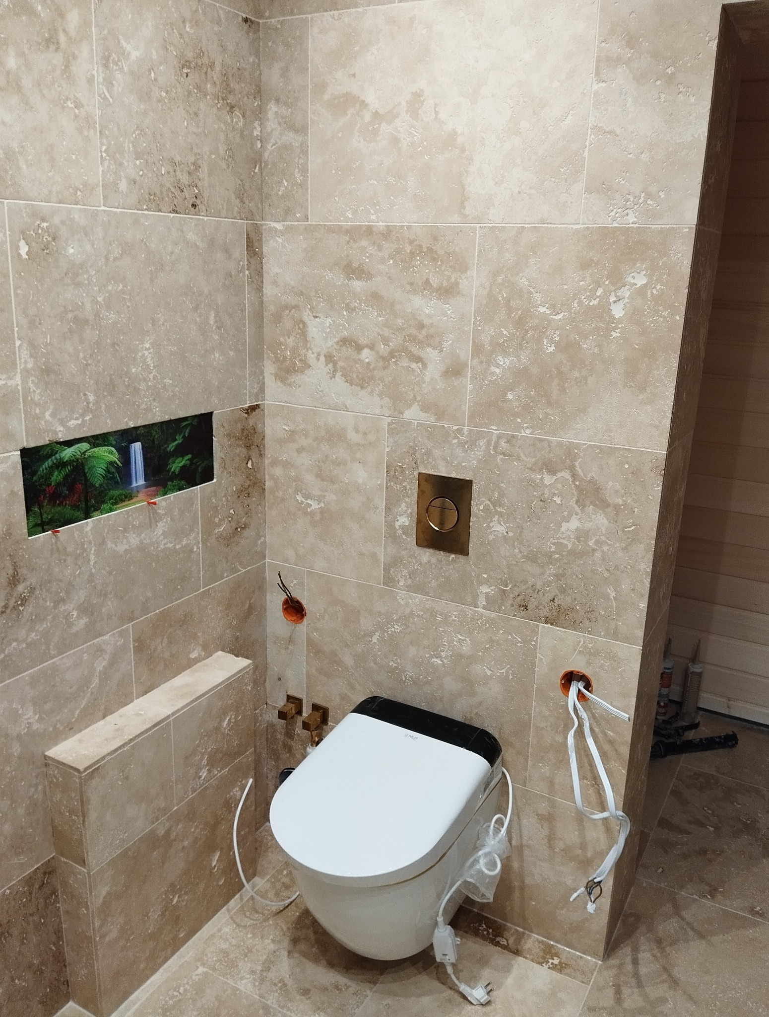 Beżowa łazienka w trakcie wykańczania: wisząca toaleta z podłączeniem elektrycznym, otwór na dekor z wodospadem, mosiężny przycisk spłuczki, widoczne kable.