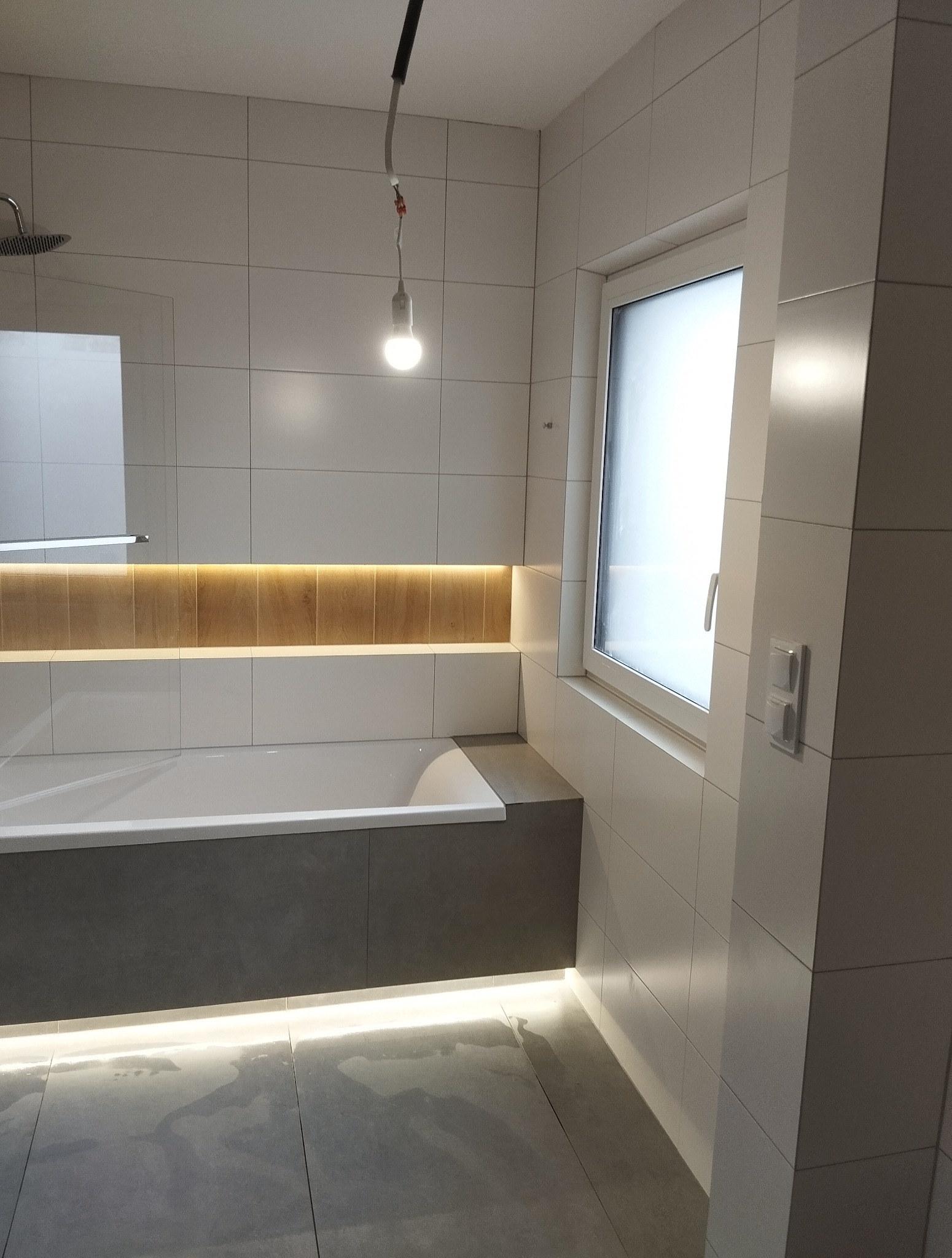 Nowoczesna łazienka z białymi płytkami, podświetlaną wanną w szarym obudowie i oknem. Minimalistyczny design z akcentami świetlnymi.