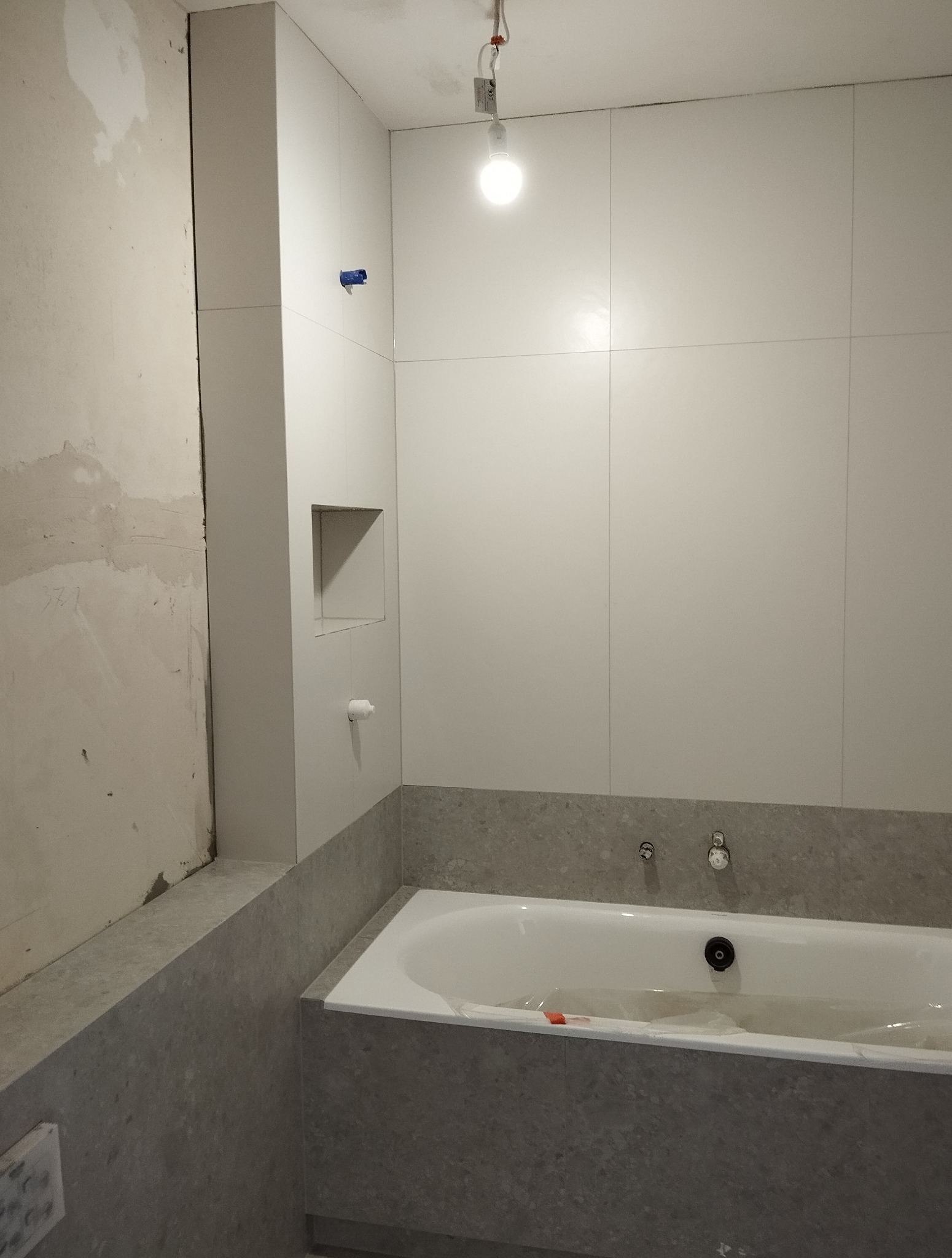 Łazienka w trakcie remontu: białe płytki, szara obudowa wanny, surowa ściana. Widoczna wnęka w ścianie i tymczasowe oświetlenie. Etap prac wykończeniowych.