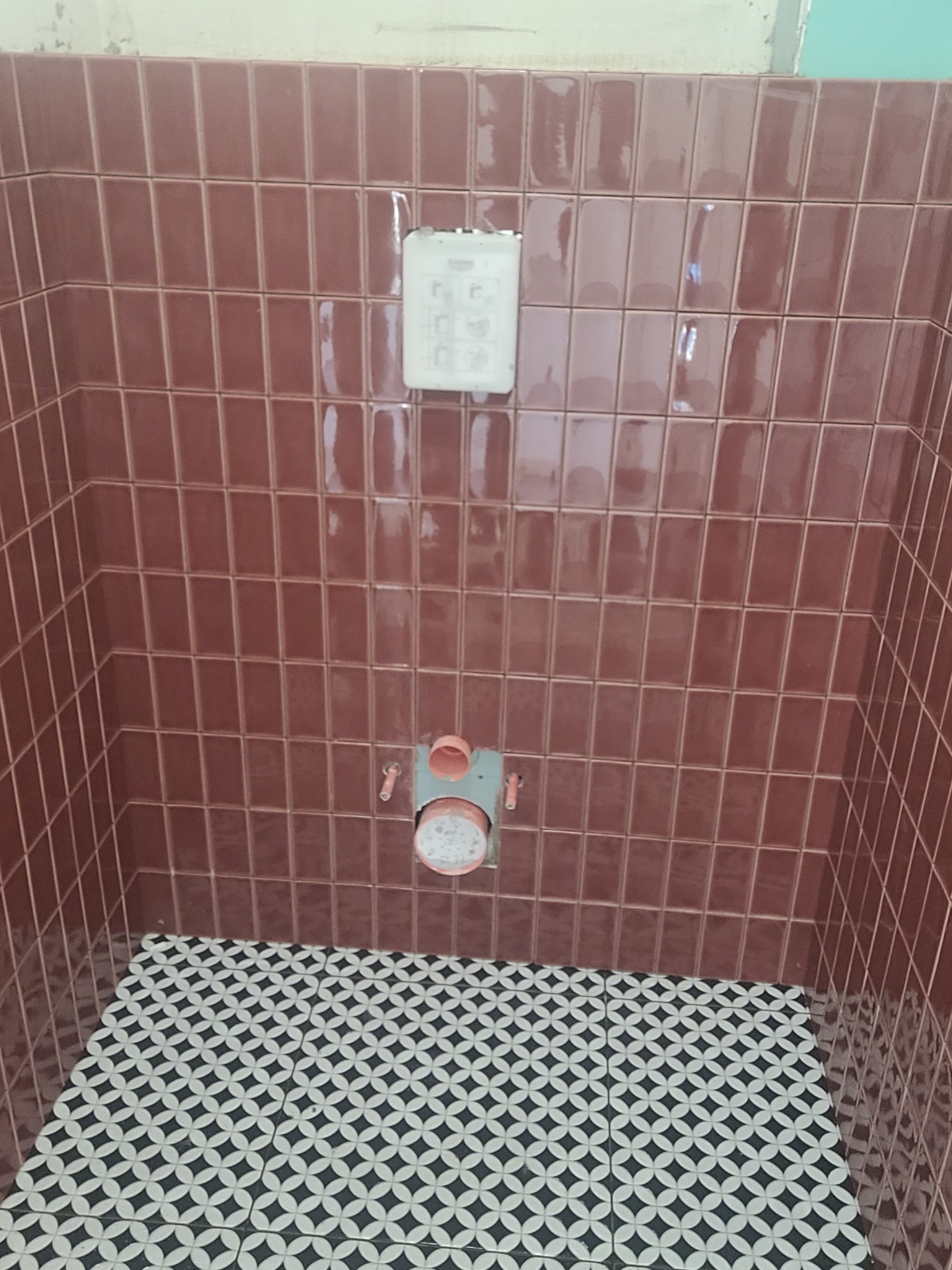 Częściowo wykończona łazienka z bordowymi, błyszczącymi płytkami na ścianach i wzorzystą, czarno-białą podłogą. Widoczne elementy instalacji wodnej.