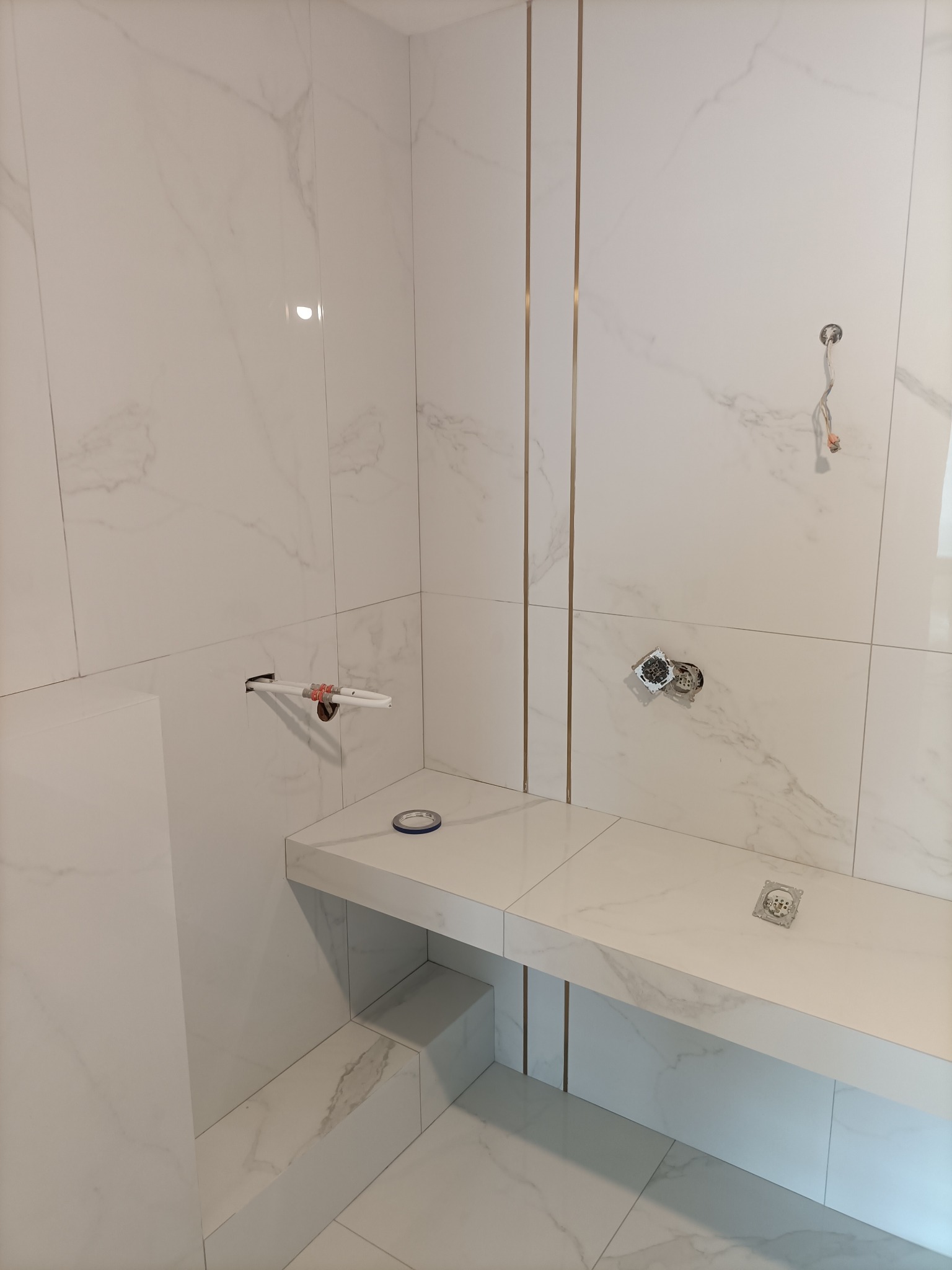 Biała łazienka z marmurowymi płytkami, widoczne przygotowane instalacje i półka wyłożona płytkami. Ujęcie z bliska, prezentujące detale wykończenia.