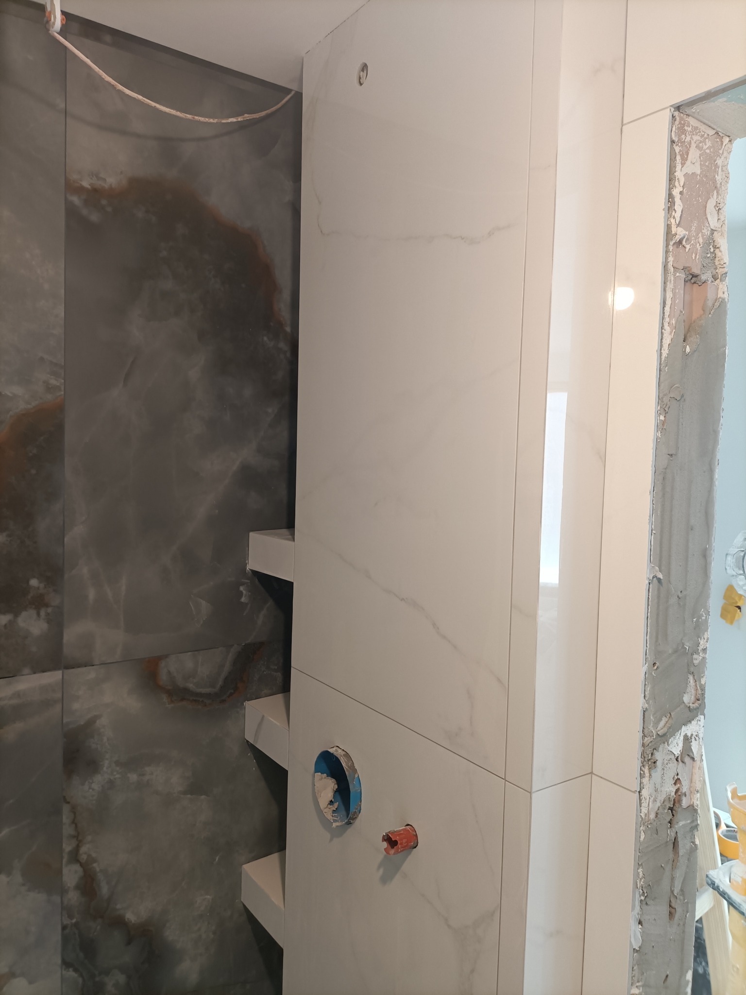 Wykończona ściana w łazience z szarymi i białymi płytkami imitującymi marmur, widoczne instalacje wodne i fragment surowego otworu drzwiowego.