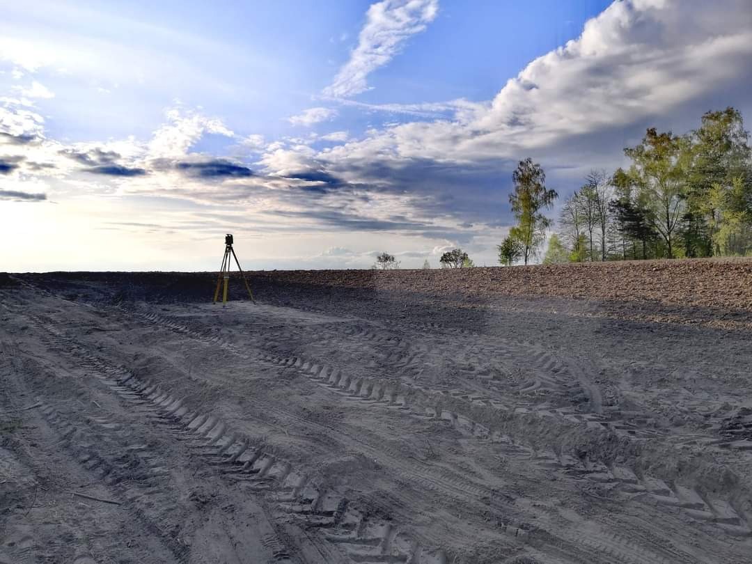 Praca geodezyjna w terenie: niwelator ustawiony na trójnogu na wyrównanym, piaszczystym terenie z widocznymi śladami opon, w tle wał ziemny z drzewami i błękitne niebo z chmurami.