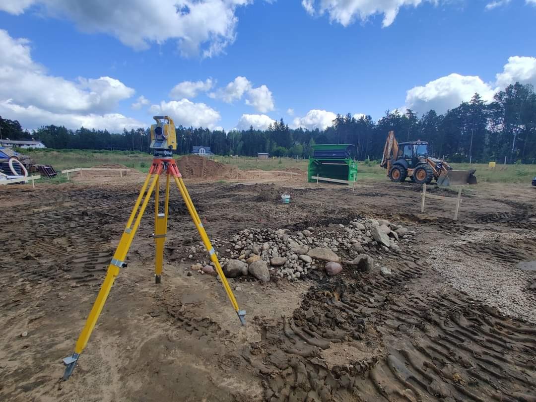 Prace przygotowawcze pod budowę w Białymstoku: żółty teodolit na trójnogu ustawiony na nierównym, błotnistym terenie, w tle widoczna koparka i przesiewacz do ziemi, błękitne niebo z białymi chmurami.