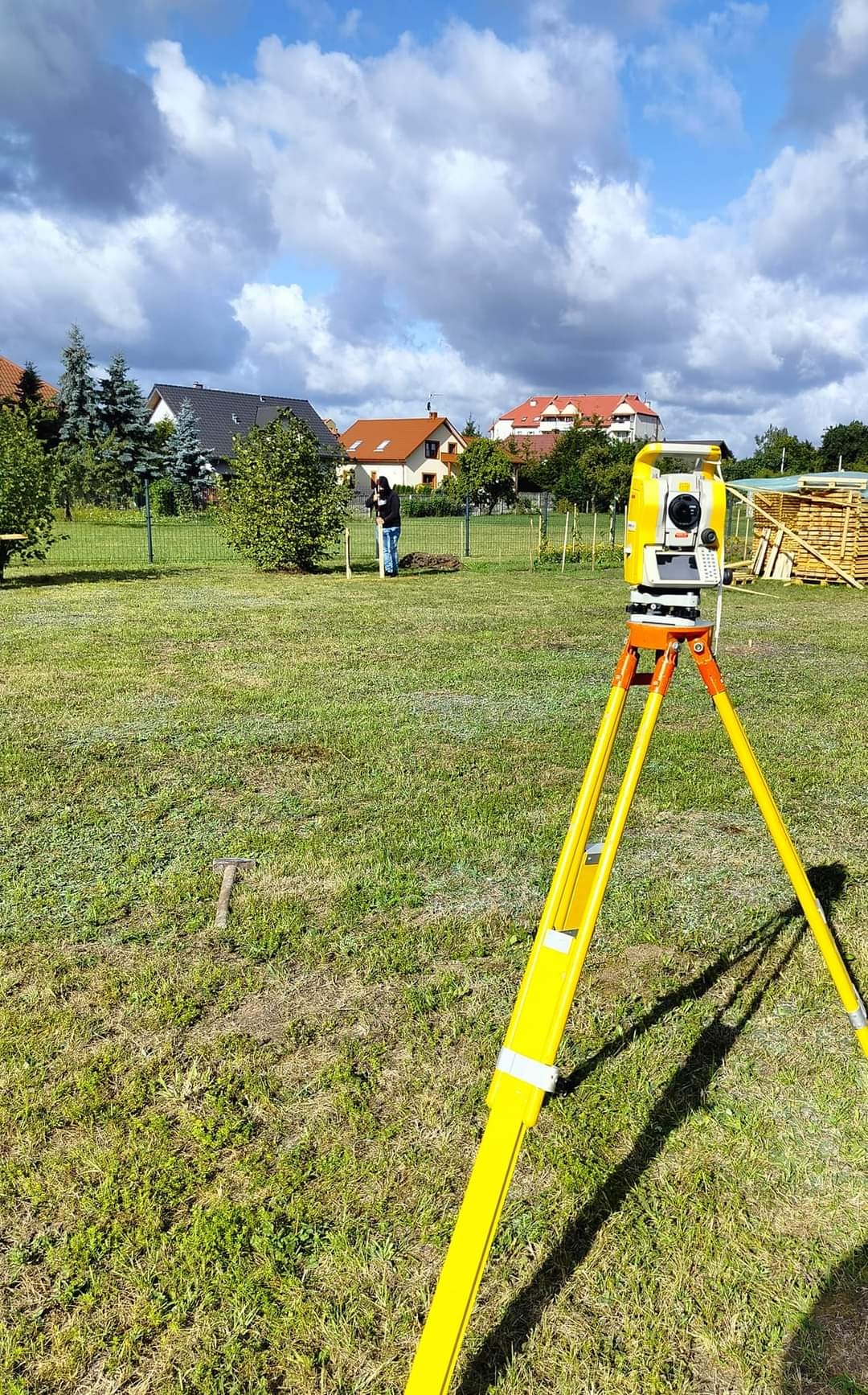Żółty teodolit ustawiony na trawie z widocznym pracownikiem w tle, wyznaczającym punkty palikami na działce budowlanej.