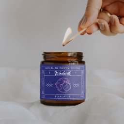 Nouva Atelier - Marta Lińska - Płonąca zapałka zbliża się do sojowej świecy o zapachu eukaliptusa w brązowym słoiku z etykietą 'Wodnik' na tle białej tkaniny.