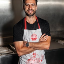 Nouva Atelier - Marta Lińska - Uśmiechnięty mężczyzna w fartuchu z logo 'Hungry Bite' na tle metalowej kuchni. Fartuch z czerwonymi ramiączkami i zabawnym nadrukiem burgera.