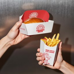 Nouva Atelier - Marta Lińska - Dwie dłonie trzymają pudełka z burgerem i frytkami z czerwonym logo 'Frenzy Bite' na białym tle. Minimalistyczny design opakowań na tle metalicznej ściany.