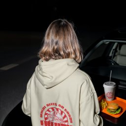 Nouva Atelier - Marta Lińska - Osoba w beżowej bluzie z nadrukiem burgera, trzymająca tacę z burgerem, frytkami i napojem na tle czarnego samochodu w nocnej scenerii.