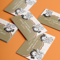 Unfold Brand - Kilka broszur 'Crème & Crust BAKED WITH LOVE' z ilustracją uśmiechniętej dziewczynki z bagietką, na beżowym tle z białym wzorem, rozłożonych na pomarańczowej powierzchni.