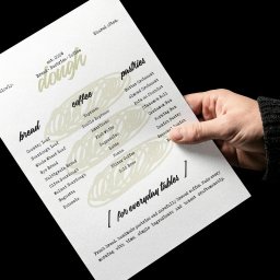 Unfold Brand - Ręka w ciemnym swetrze trzyma białą kartkę z menu 'dough' (est. 2026) o minimalistycznym designie, prezentującą listę pieczywa, kaw i wypieków, m.in. chleb na zakwasie i croissanty.