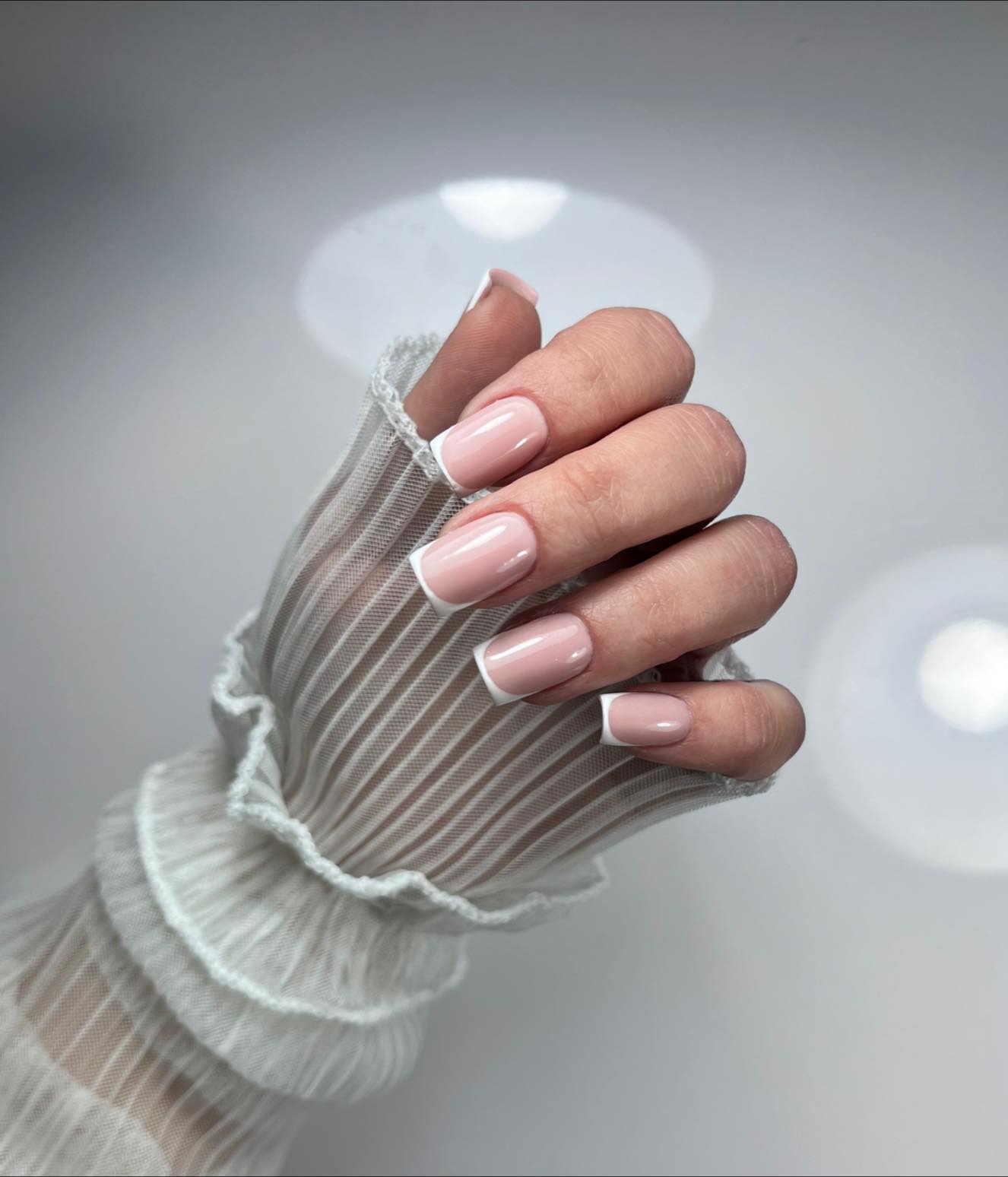 Dłoń w przezroczystej rękawiczce z falbanką prezentuje świeżo wykonany manicure francuski na krótkich, kwadratowych paznokciach w odcieniach różu i bieli.