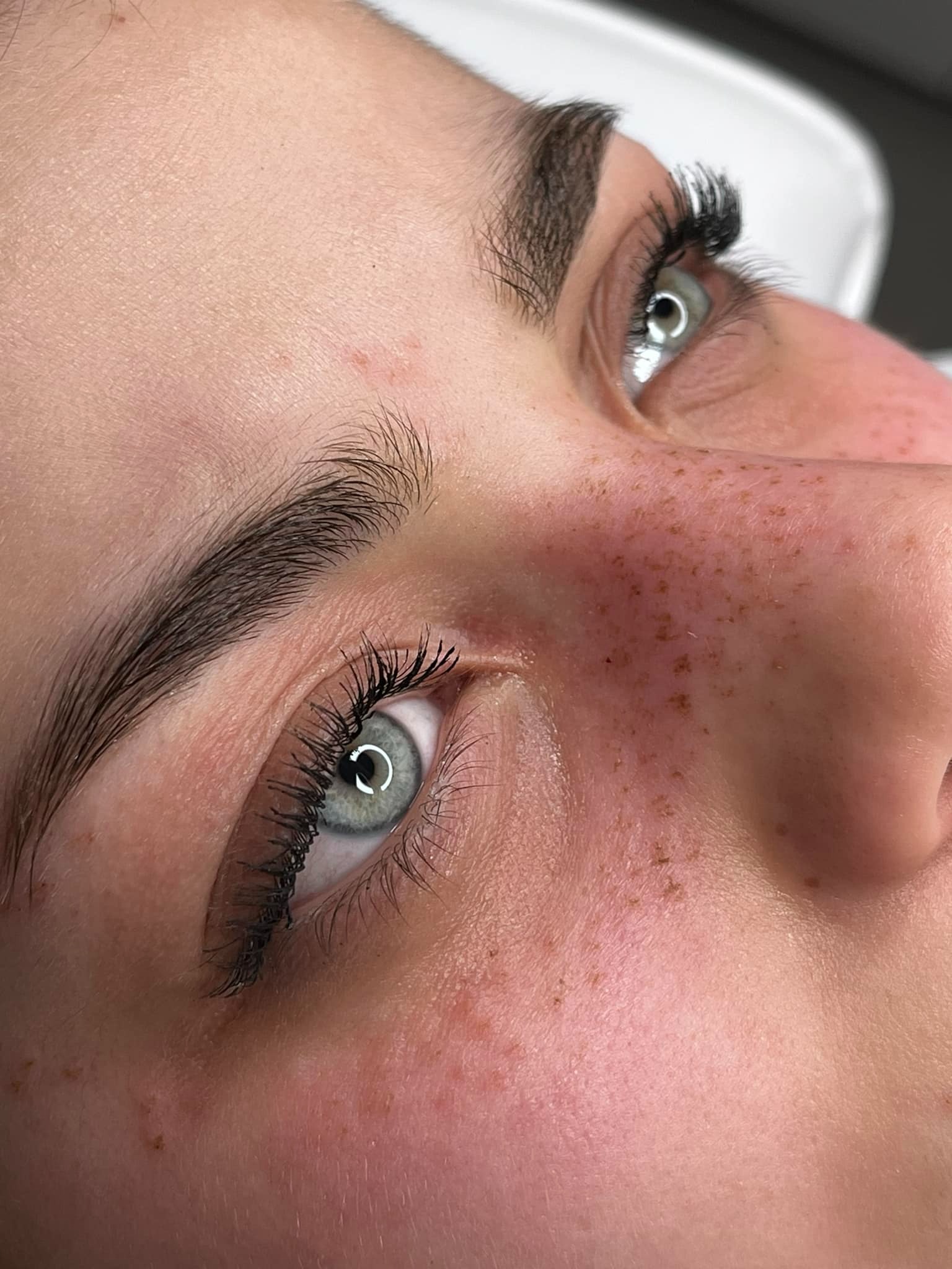 Zbliżenie na twarz kobiety z widocznymi, świeżo przedłużonymi rzęsami i brwiami po zabiegu laminacji, z delikatnymi piegami na policzkach, uchwycone pod kątem z góry.