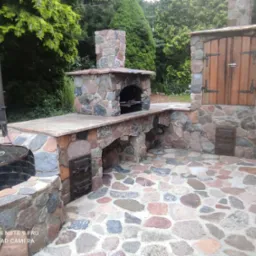 Kamienna zabudowa grilla ogrodowego z kominem i szafką, wykończona kostką brukową z kamienia polnego, na tle zieleni.