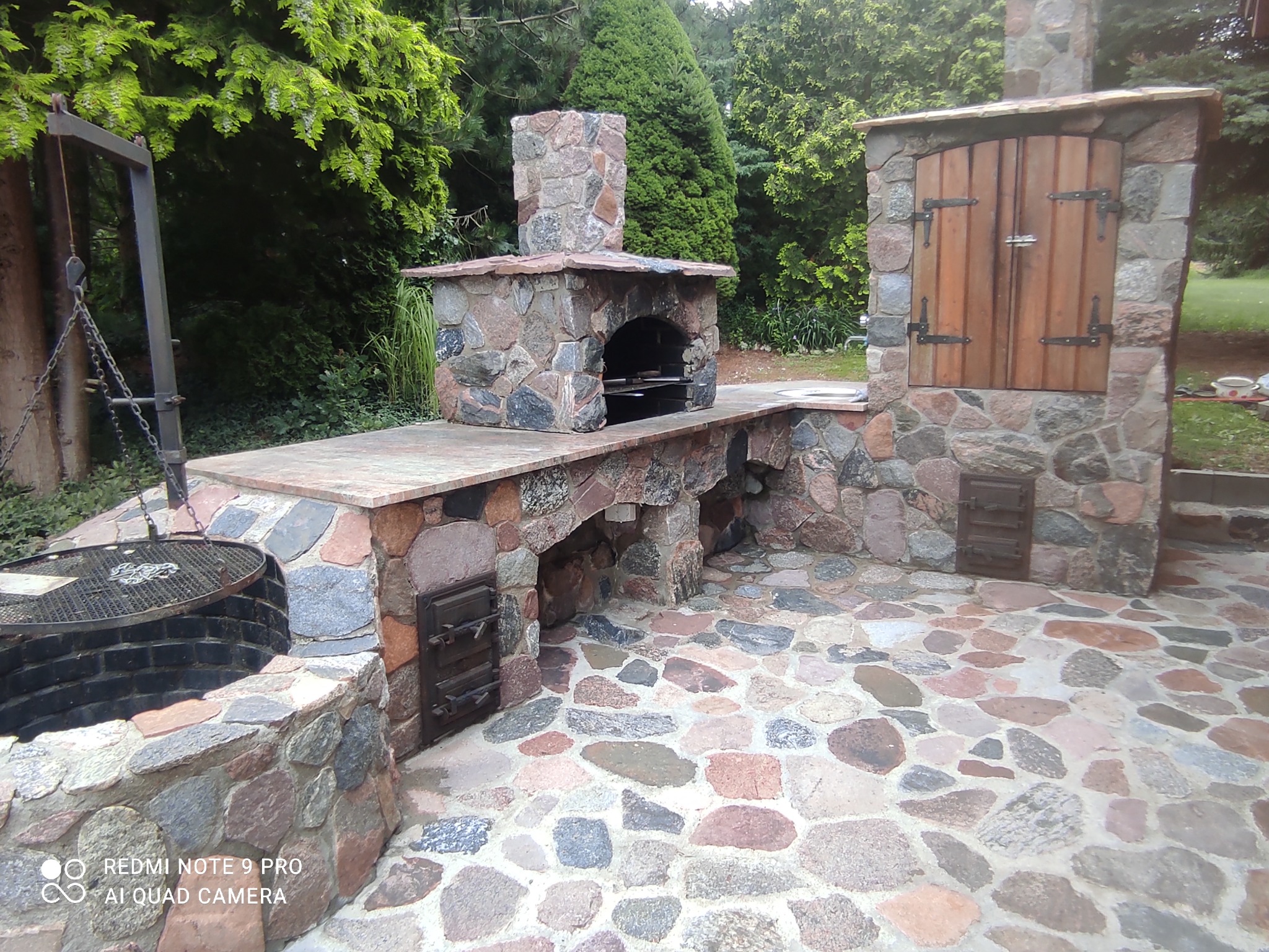 Kamienna zabudowa grilla ogrodowego z kominem i szafką, wykończona kostką brukową z kamienia polnego, na tle zieleni.