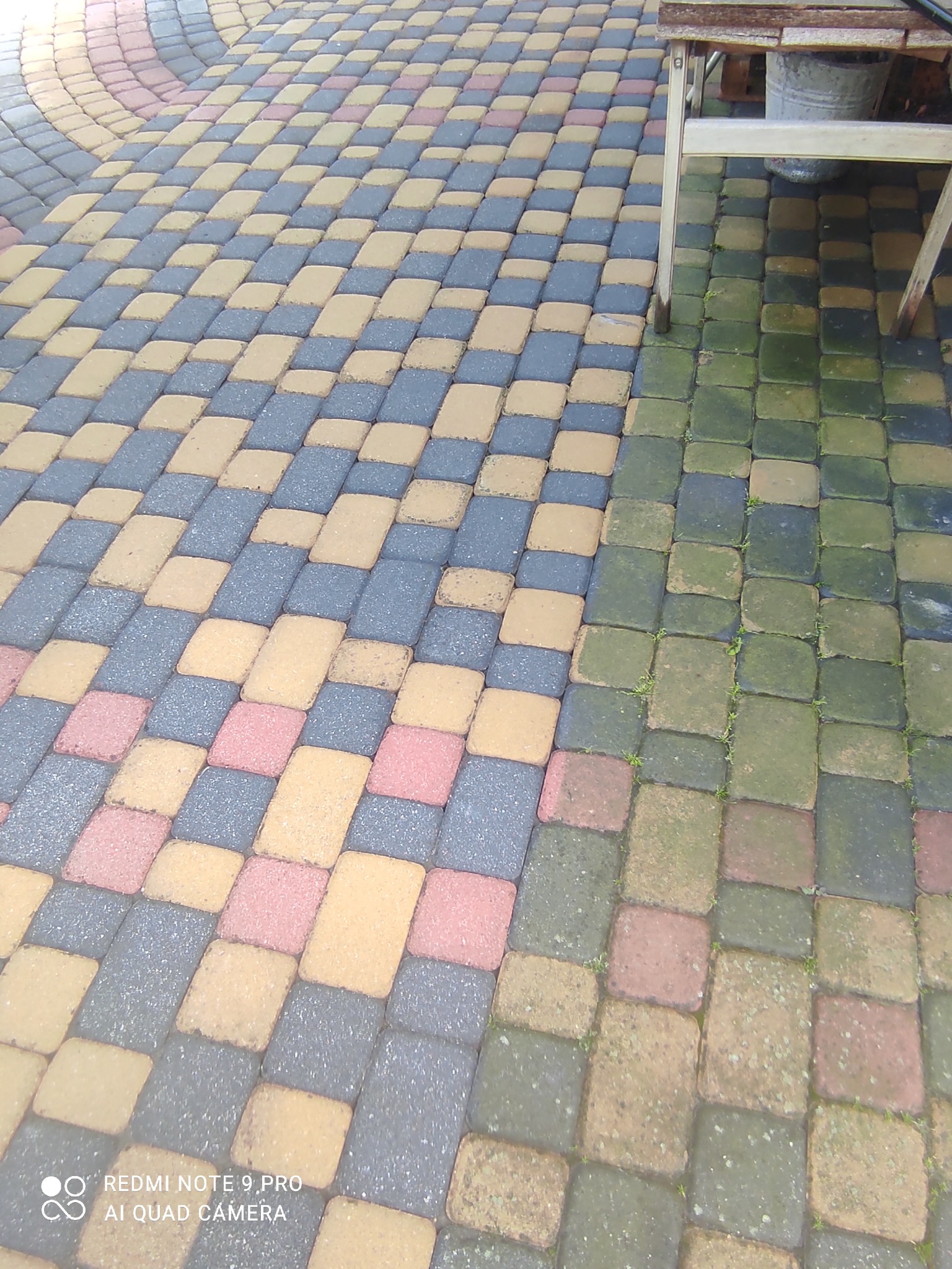 Kolorowa kostka brukowa ułożona we wzór, część wyczyszczona, a część pokryta mchem i zabrudzeniami, obok stoi stolik.