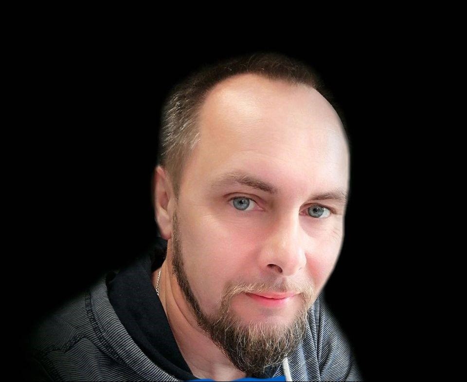 Portret mężczyzny z lekkim zarostem, niebieskimi oczami i szaro-czarną bluzą na czarnym tle.