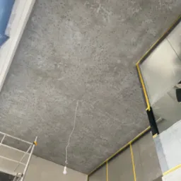 Szary sufit z efektem dekoracyjnym, zabezpieczone folią ściany, widoczny fragment rusztowania i wisząca żarówka.