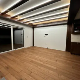 Wykończone wnętrze z drewnianą podłogą, przesuwnymi drzwiami balkonowymi i sufitem podwieszanym z oświetleniem LED, widoczne przygotowane punkty instalacji elektrycznej na ścianie.