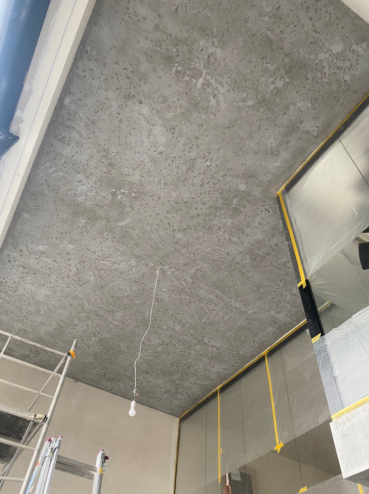 Szary sufit z efektem dekoracyjnym, zabezpieczone folią ściany, widoczny fragment rusztowania i wisząca żarówka.