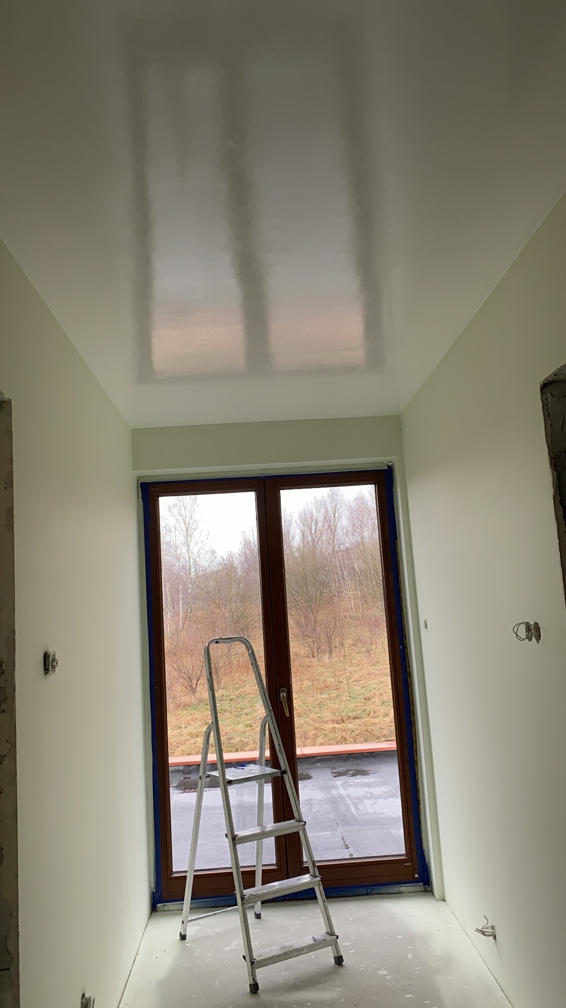Świeżo pomalowany sufit w jasnym odcieniu z widocznymi refleksami światła, drabina aluminiowa oparta o okno balkonowe zabezpieczone folią malarską, widok na zewnątrz z drzewami bez liści.