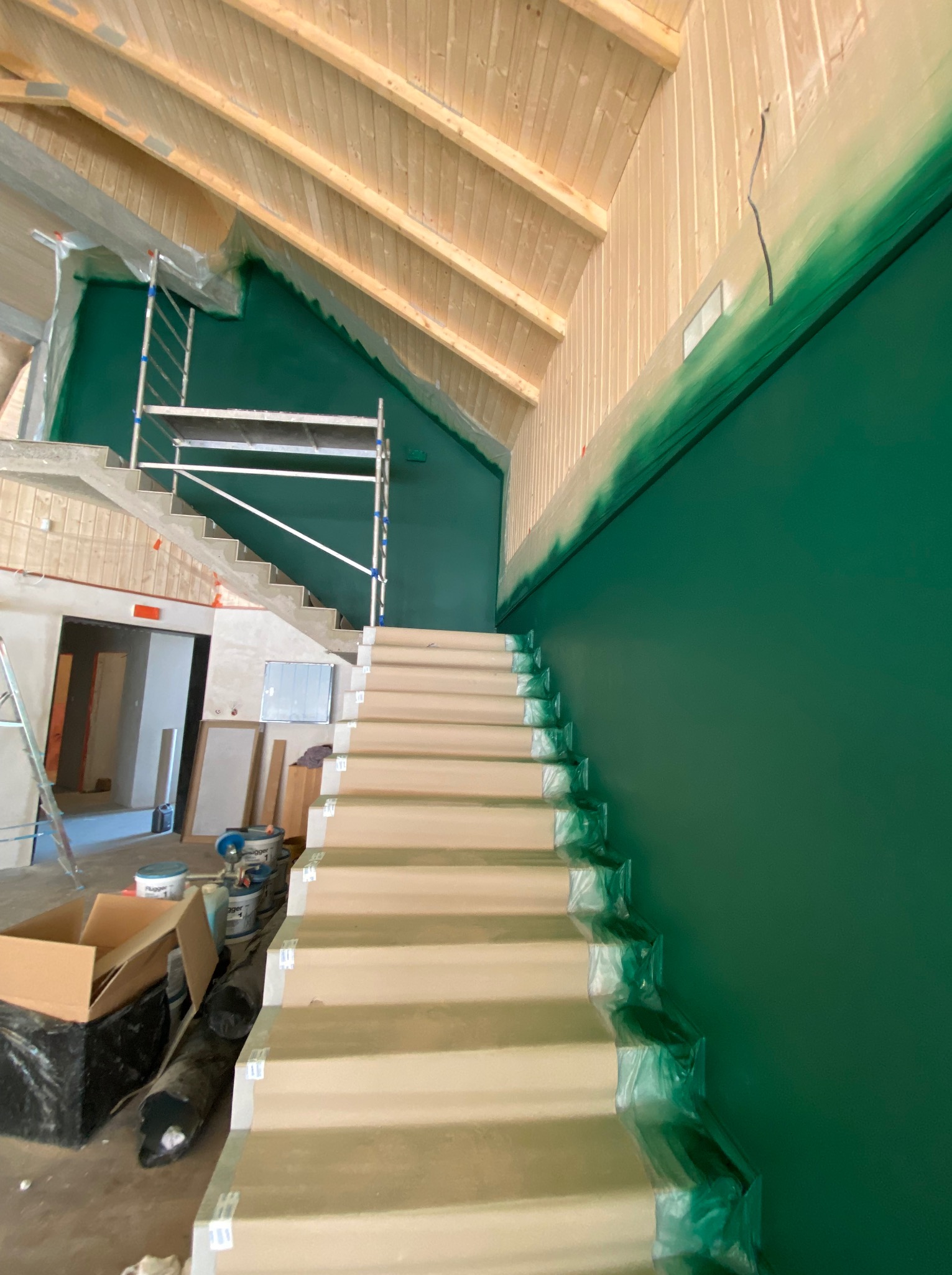 Malowanie ścian na intensywny zielony kolor w dwukondygnacyjnym pomieszczeniu ze schodami, zabezpieczonymi folią malarską, widoczne rusztowanie i drewniane wykończenie poddasza.