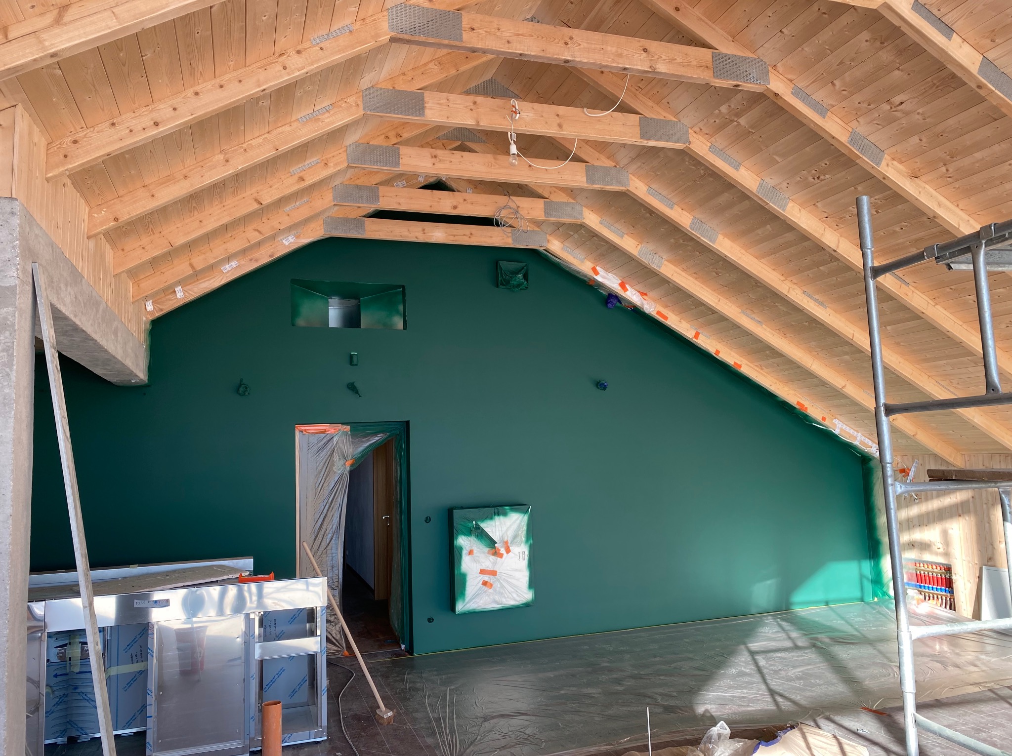 Wnętrze w trakcie prac wykończeniowych: ściana pomalowana na ciemnozielony kolor, drewniany szkielet dachu z widocznymi metalowymi łącznikami, podłoga zabezpieczona folią, widoczne elementy...