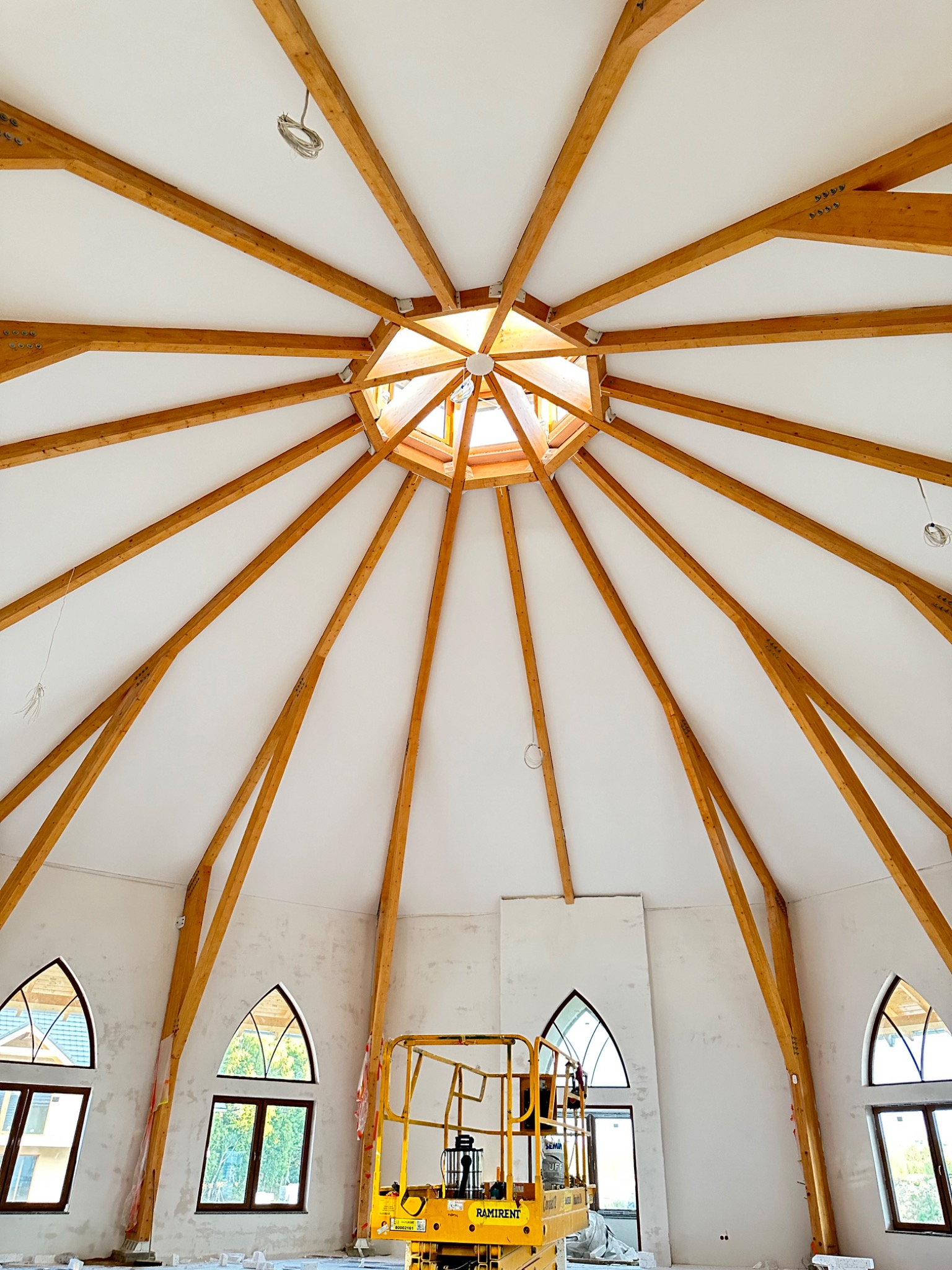 Wnętrze okrągłego budynku z drewnianą konstrukcją dachu w kształcie gwiazdy, białe ściany w trakcie prac wykończeniowych, podnośnik koszowy w kolorze żółtym i gotyckie okna.