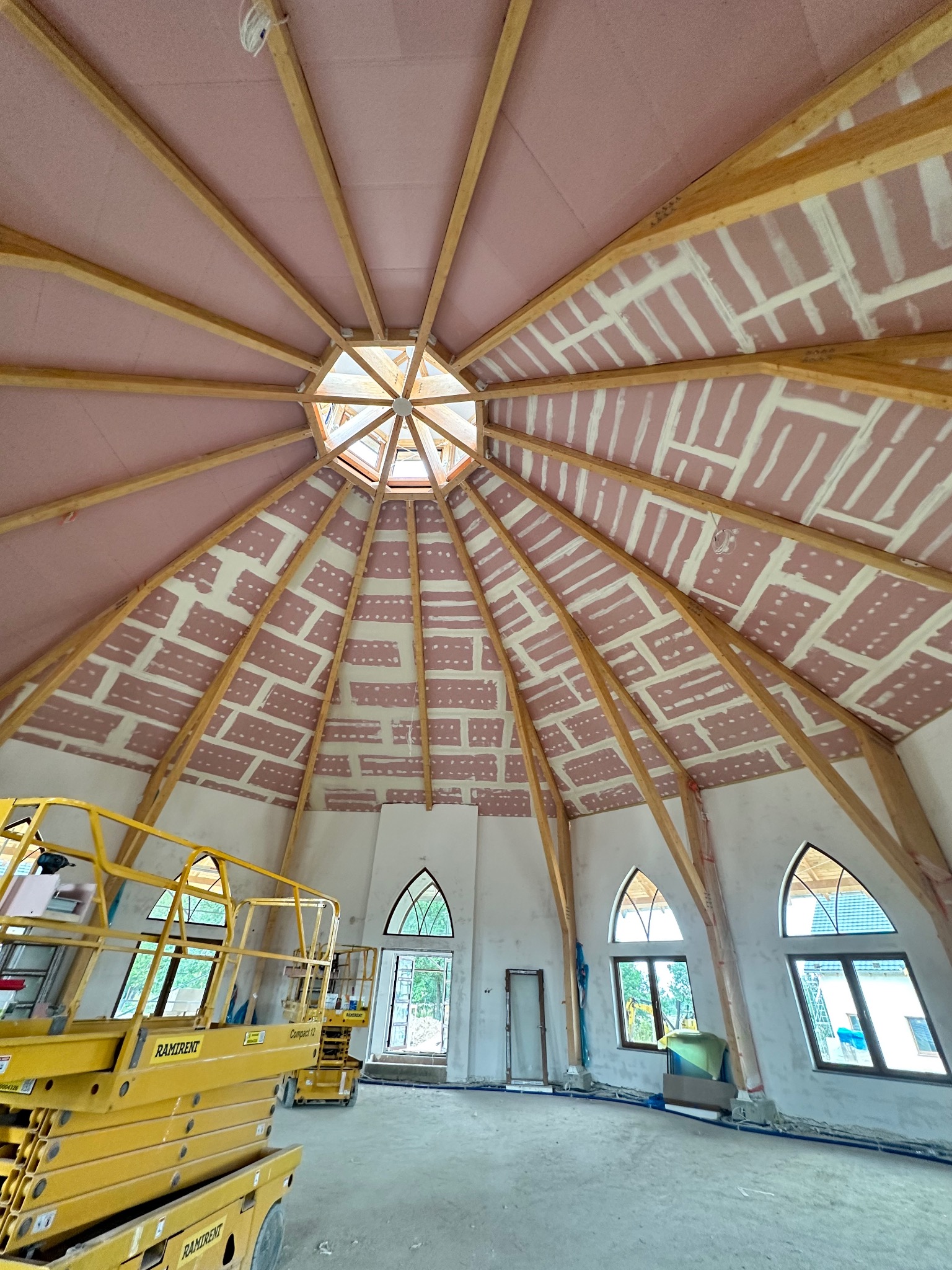 Wnętrze kopuły w trakcie budowy: widoczne drewniane elementy konstrukcyjne, różowa izolacja, fragmenty białej ściany, okna w kształcie łuku oraz żółty podnośnik koszowy.