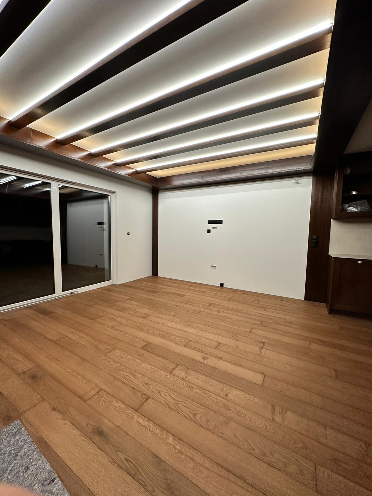 Wykończone wnętrze z drewnianą podłogą, przesuwnymi drzwiami balkonowymi i sufitem podwieszanym z oświetleniem LED, widoczne przygotowane punkty instalacji elektrycznej na ścianie.