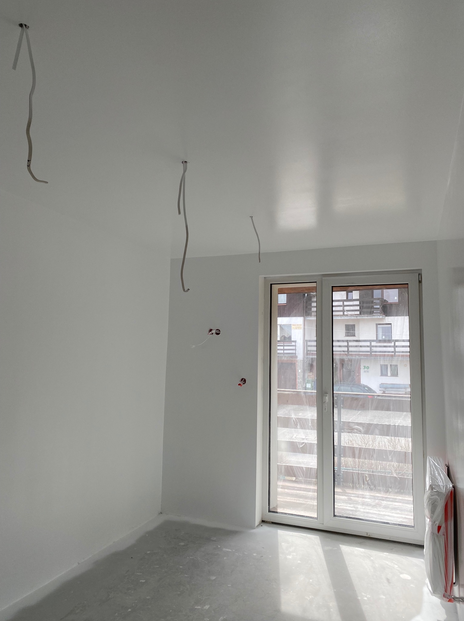 Wnętrze pokoju po remoncie z gładkimi, białymi ścianami i sufitem, widoczne wystające kable elektryczne i okno balkonowe z widokiem na budynek mieszkalny.