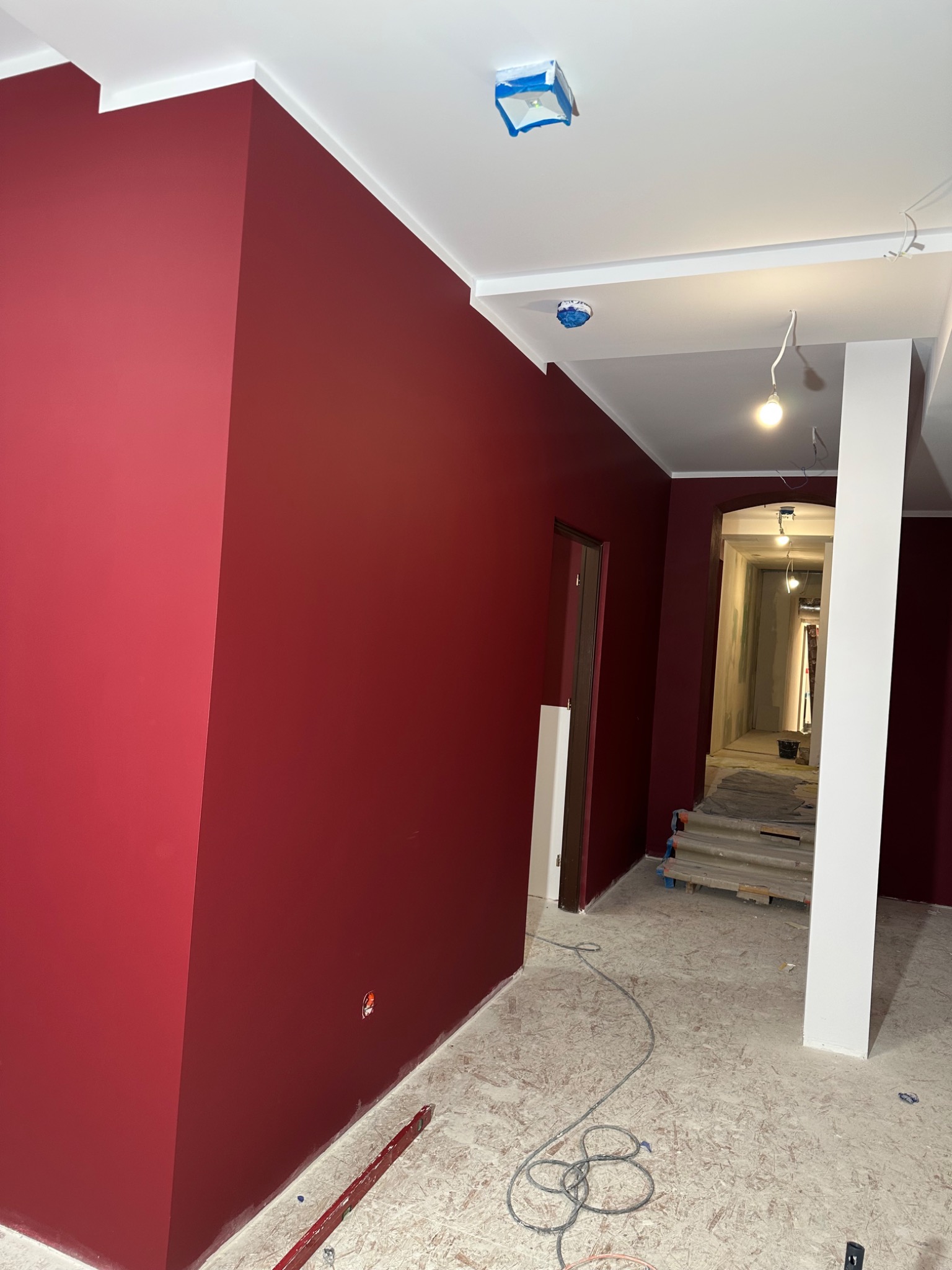 Świeżo pomalowane na bordowo ściany korytarza z białymi listwami sufitowymi i kolumną, widoczne zabezpieczone folią lampy sufitowe, nieukończona podłoga i narzędzia w tle.