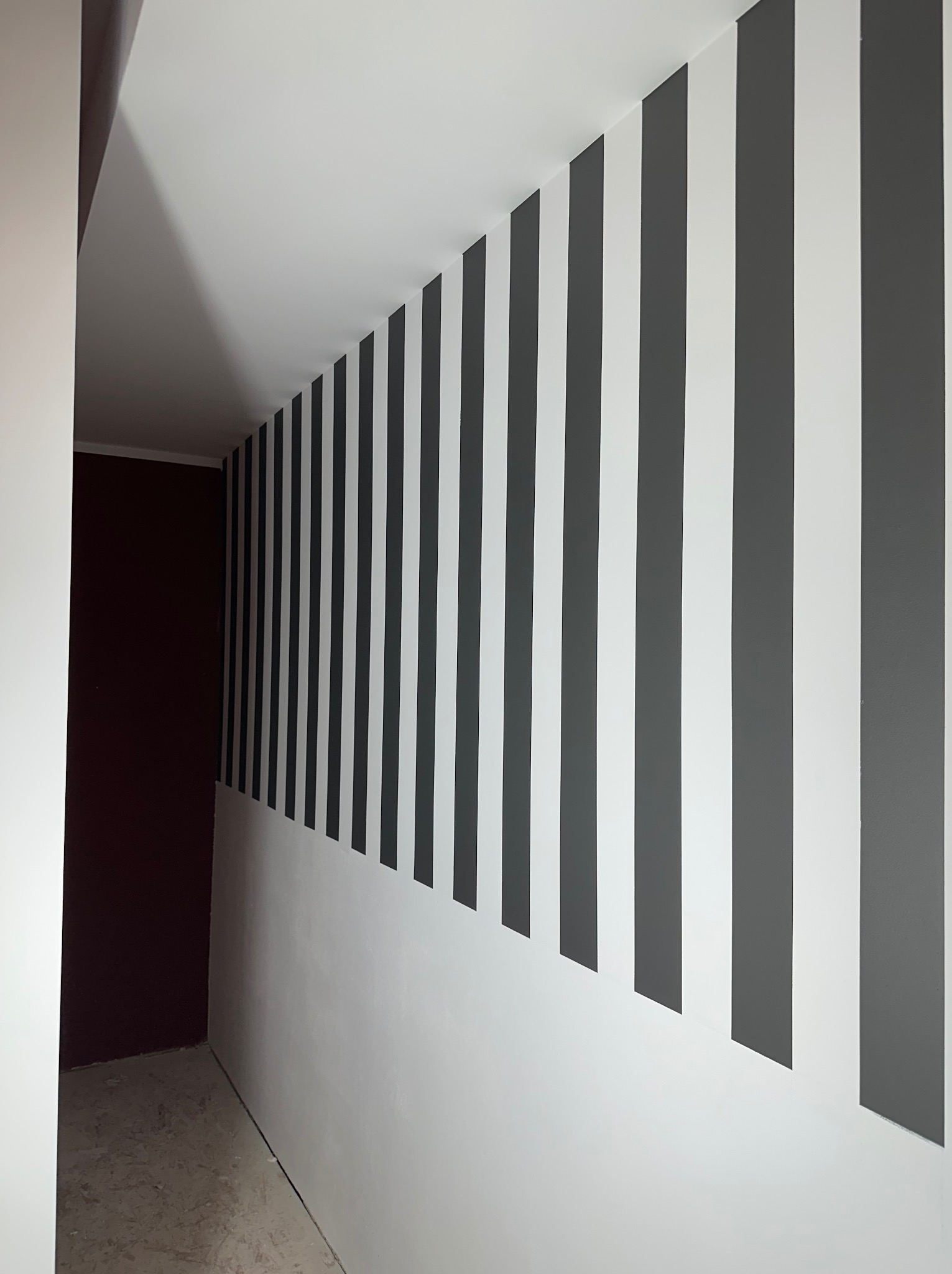Korytarz z pomalowaną ścianą w pionowe, szaro-białe pasy, widoczny fragment ciemnej ściany po lewej stronie, surowa podłoga w tle.