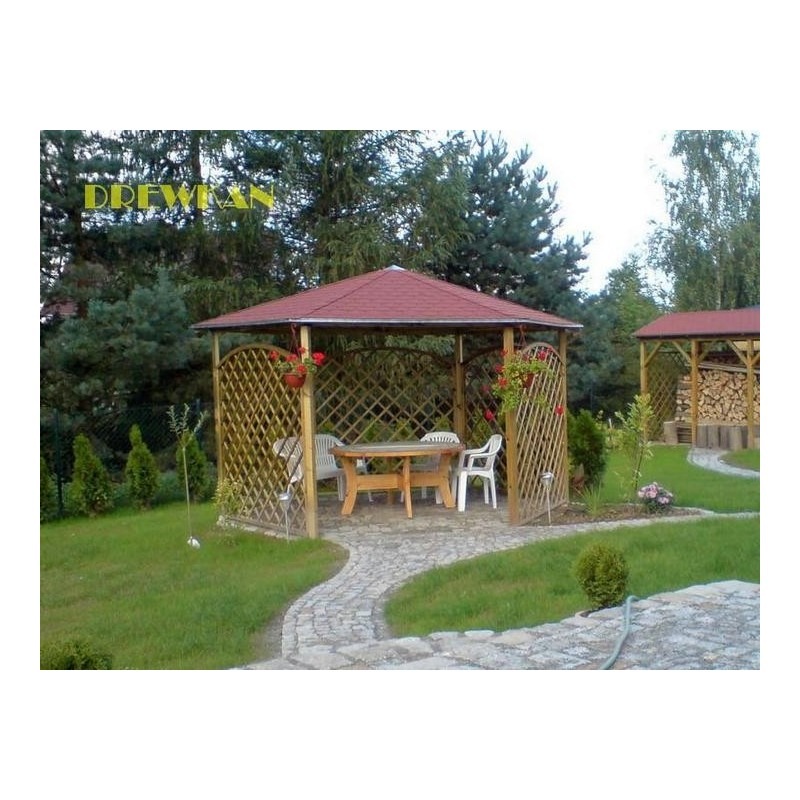 Drewniana konstrukcja z czerwonym dachem, ażurowe ściany, stół i krzesła wewnątrz, donice z kwiatami, kręta ścieżka z kamienia, w tle widoczna druga podobna konstrukcja z ułożonym drewnem.