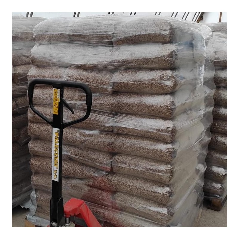 Stos worków z pelletem drzewnym, zabezpieczonych folią, na palecie, widoczne uchwyty wózka paletowego.