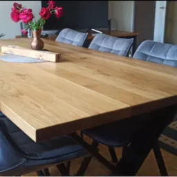 Jasny drewniany stół jadalny z blatem z litego drewna, otoczony szarymi krzesłami z welurowym obiciem, na stole dekoracje: wazon z czerwonymi różami i drewniany pojemnik.