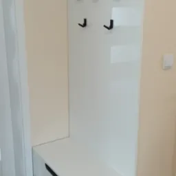 Biała szafka z dwiema szufladami i panelem z wieszakami w przedpokoju, czarne uchwyty i wieszaki, minimalistyczny design.