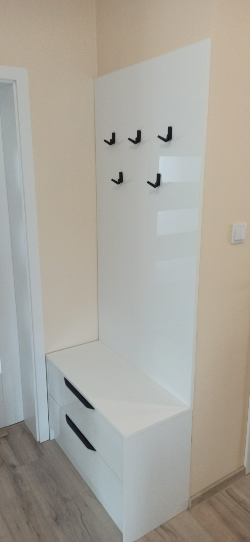 Biała szafka z dwiema szufladami i panelem z wieszakami w przedpokoju, czarne uchwyty i wieszaki, minimalistyczny design.