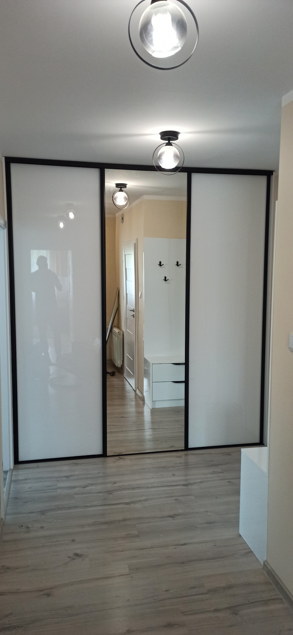 Szafa przesuwna z białymi frontami i czarnymi ramami w przedpokoju, widoczne odbicie osoby w lustrzanych drzwiach, oświetlenie sufitowe.