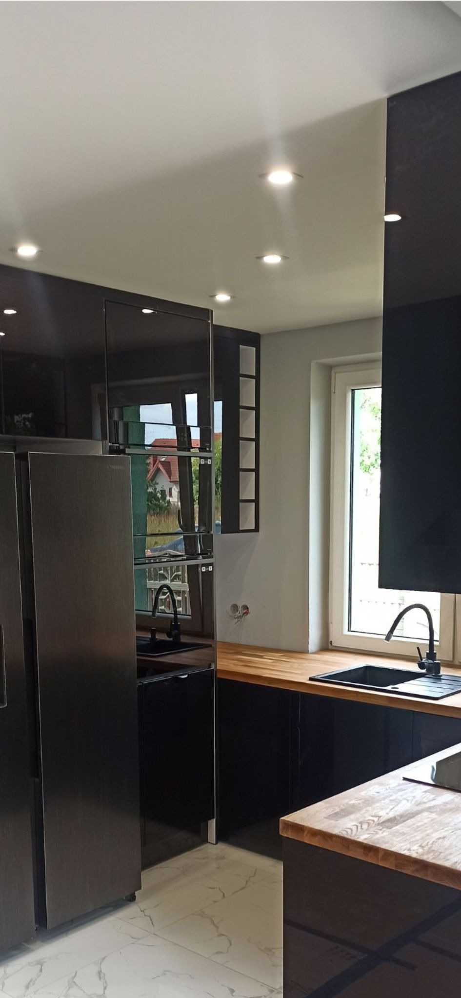 Nowoczesna, czarna kuchnia z drewnianym blatem, widok na zewnątrz przez okno, marmurowe płytki podłogowe i oświetlenie punktowe.