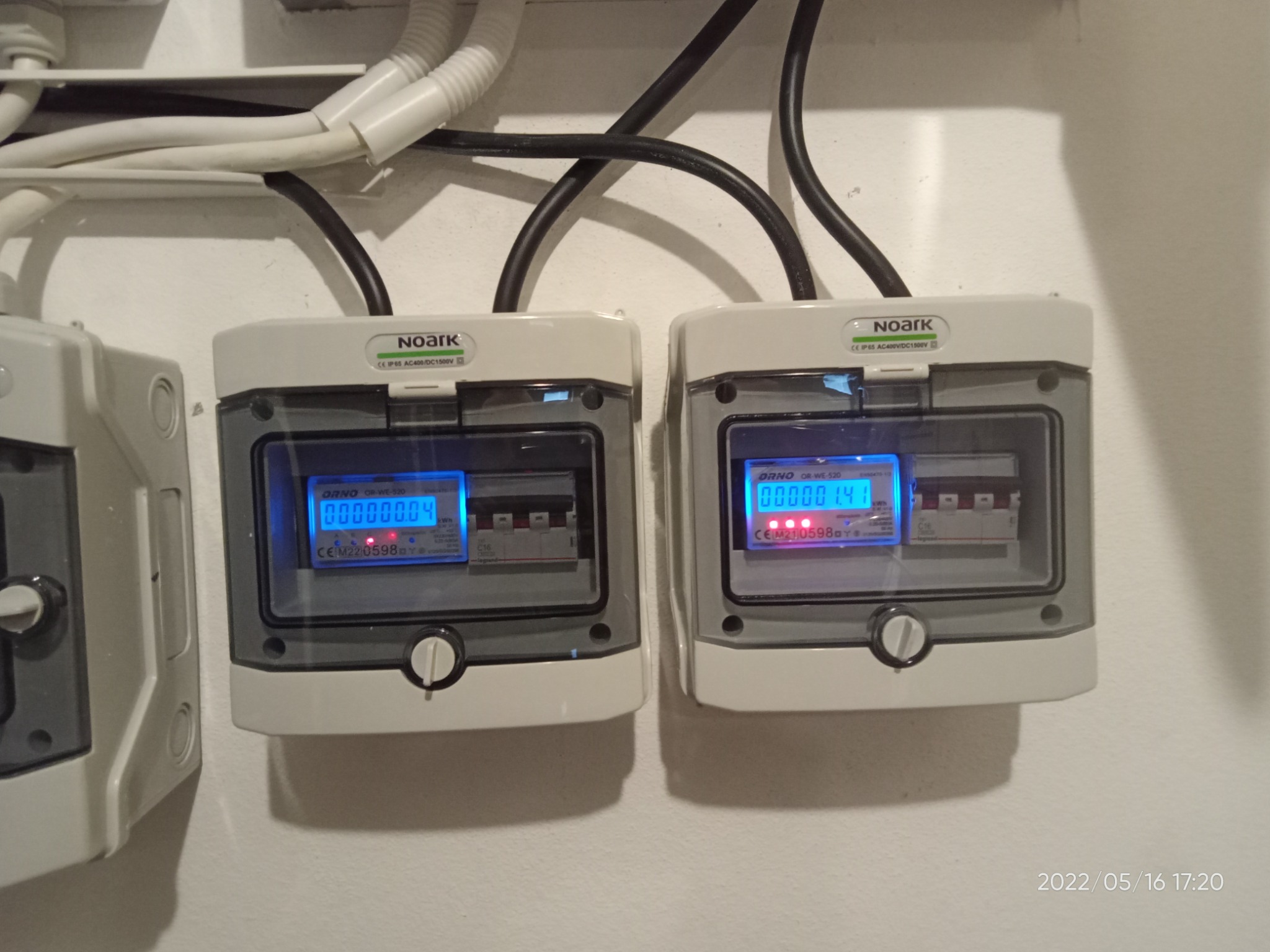 Dwa liczniki energii elektrycznej marki Noark z wyświetlaczami LED wskazującymi zużycie prądu, zamontowane na ścianie z widocznymi kablami w peszlach.